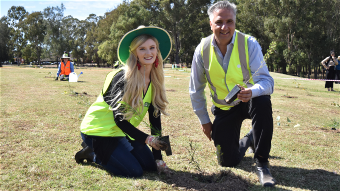 Mayor Frank Carbone and Miss Earth Australia Brittany Dickson, wearing high visibility vests, smiling and posing while planting tree saplings 