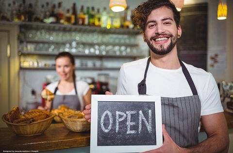 Two bakery employees smiling at camera with one holding up a chalkboard with the word open written on it