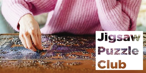 A woman focused on assembling a jigsaw puzzle on a wooden table, surrounded by scattered puzzle pieces.