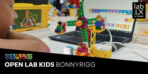 A child sits at a desk with a laptop open, playing LEGO toy car beside them.